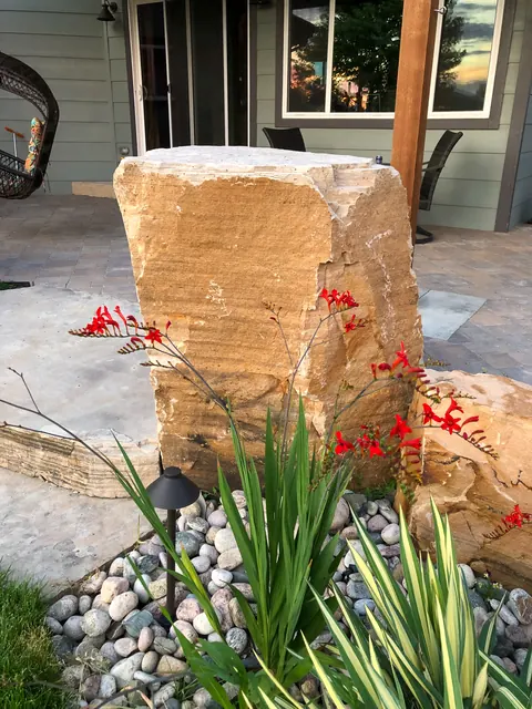 Standing sandstone boulder with red crocosmia, ornamental grass and a path light at a patio edge.