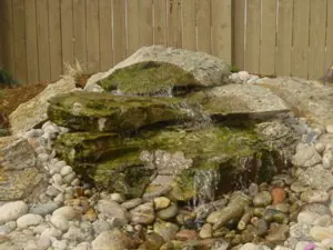 Small bubbling boulder fountain with moss and river rock in front of a wood fence.