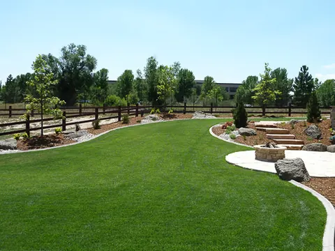 Curving lawn leads to a flagstone patio with a small fire pit along a rail fence.