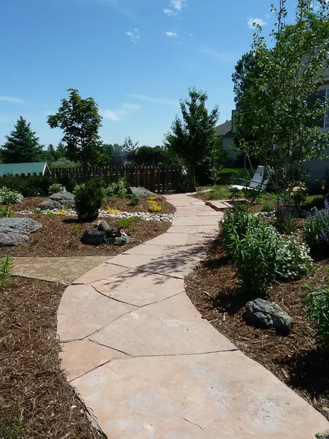 Curving buff flagstone walkway through a garden of shrubs, boulders and mulch.