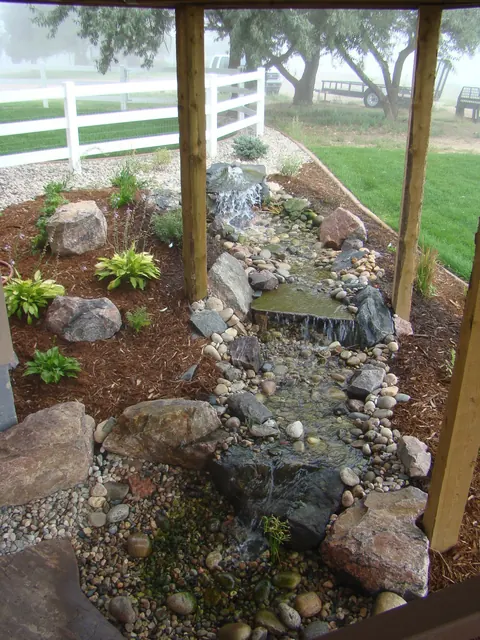 Narrow dry stream bed of river rock and boulders winding beneath a wooden deck.