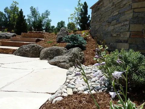 Flagstone path beside boulders, hosta and river rock below a stacked stone wall and steps.