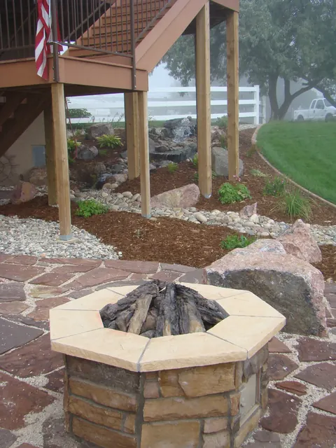Red flagstone patio with a hexagonal stone fire pit and boulders beneath a deck.