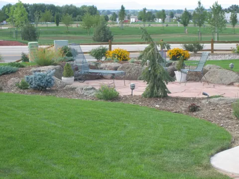 Flagstone patio with chairs, weeping evergreen and boulders beside a curved lawn.