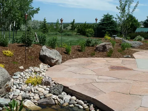 Red flagstone patio edged with river rock and boulders overlooking a golf-course style open lawn.