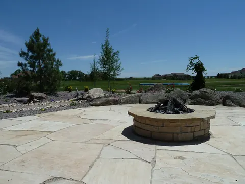Large buff flagstone patio with a round stone fire pit and boulders beneath open sky.