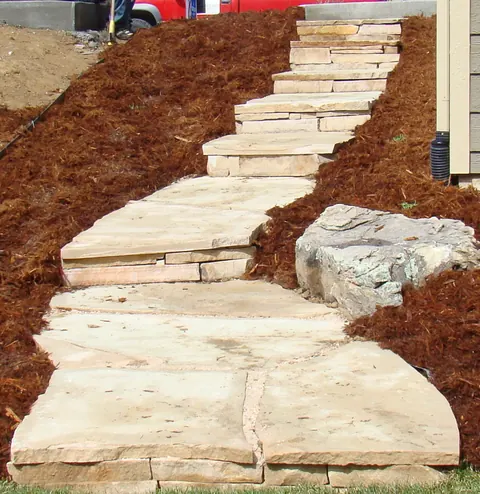 Buff flagstone slab steps climbing a mulched hillside beside a boulder.