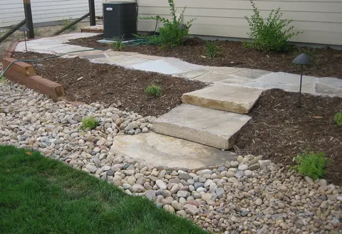 Flagstone stepping path with wide slab steps, mulch, shrubs and river rock beside a lawn.