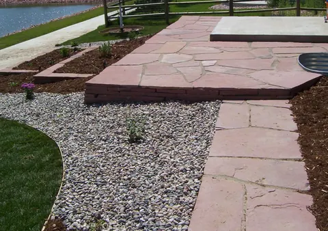 Red flagstone walkway to a raised flagstone landing with gravel beds and a pond view.