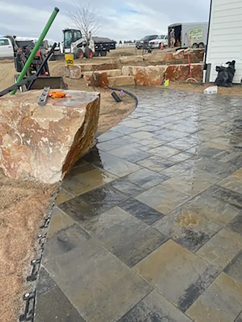 Dark paver patio under construction beside a sandstone boulder and stacked rock steps in Timnath.