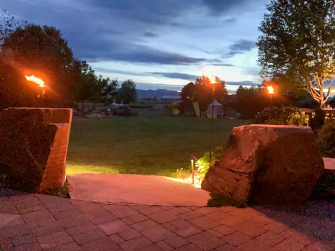 Brick paver patio at dusk with tall boulders, path lights and flaming torches overlooking a lawn.