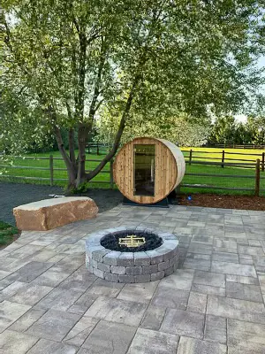 Gray paver patio with a stone ring fire pit and a barrel sauna beneath shade trees.