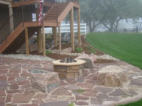 Red flagstone patio with a stone fire pit and boulders beneath a wooden deck.
