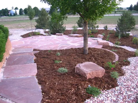 Red flagstone walkway and boulder beside a curved mulch bed under a young shade tree.