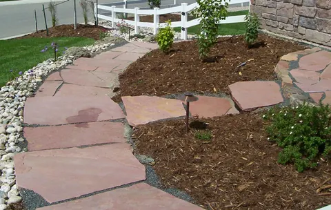 Red flagstone path with a low-voltage path light, mulch beds and a white fence.