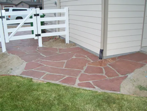 Red flagstone patio beside a white vinyl gate and lawn at the side of a home.
