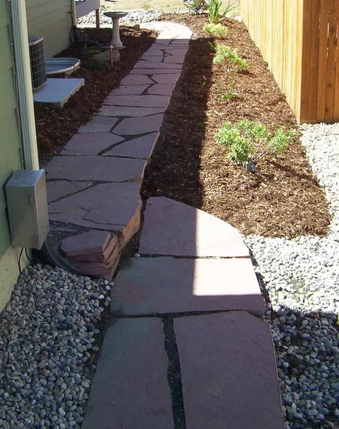 Narrow red flagstone path in a side yard with river rock, mulch and a wood fence.