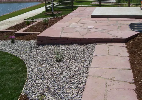 Red flagstone walkway leading to a raised flagstone landing with gravel beds beside a pond.