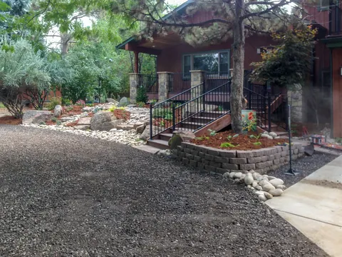 Block retaining wall with stairs and iron railing beside a gravel drive and river rock bed.