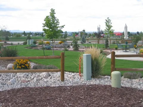 River rock and mulch bed with grasses and yellow flowers along a split rail fence.