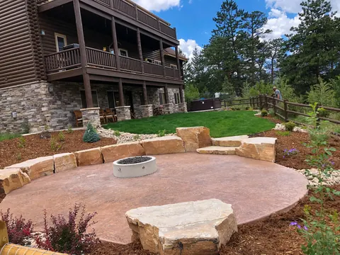 Round stamped concrete patio with a fire pit and flagstone boulder seating beside a mountain-style home.