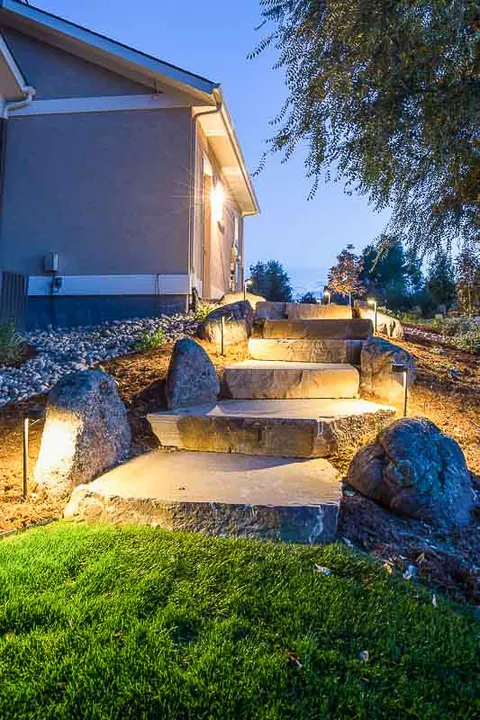 Boulder steps lit by low-voltage path lights at dusk beside a lawn and stucco home.