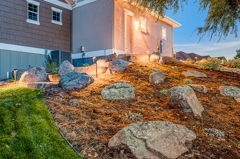 Landscape lights glowing on a hillside of granite boulders and mulch beside a home.