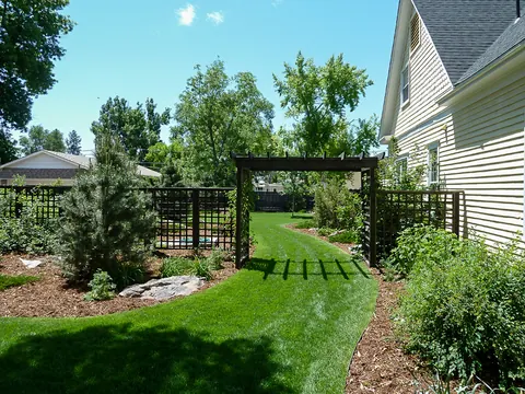 Backyard lawn with a trellis arbor, evergreens and mulch beds along the side of a home.
