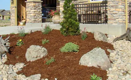 Two boulders and small plants in a mulch bed with river rock edging below a porch.