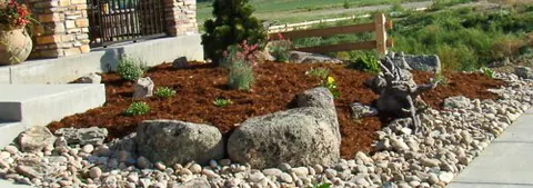 Mulch bed with large boulders and river rock edging along a sidewalk beside a stone porch.