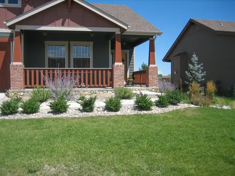 Foundation plantings of small shrubs in river rock below a craftsman porch with a green lawn.