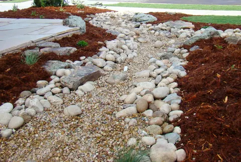 Winding dry creek bed of river rock and gravel through red mulch beside a flagstone patio.
