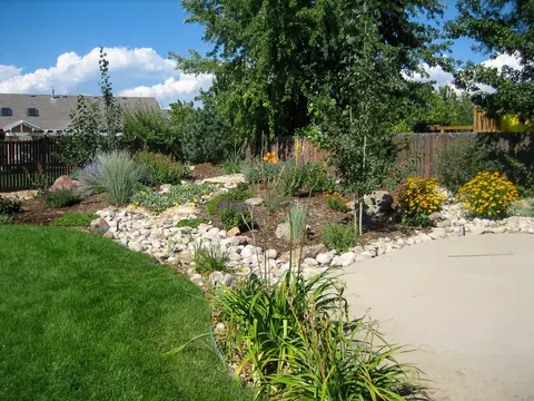 River rock dry creek with daylilies and yellow flowers beside a lawn and concrete path.