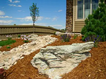 Dry creek of white river rock around a limestone boulder in a mulch bed.