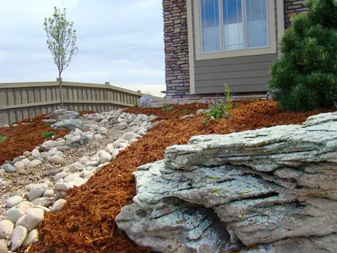Dry creek of river rock curving past a weathered limestone boulder along a wood fence.