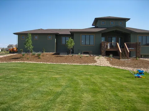 Green craftsman home with a large lawn and mulch and gravel planting beds with young trees.