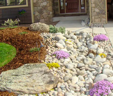 River rock bed with purple creeping phlox, yellow flowers and a flat boulder by an entry.