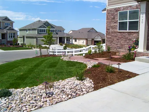 River rock dry creek beside a sidewalk, mulch bed and lawn at a brick-accented home.