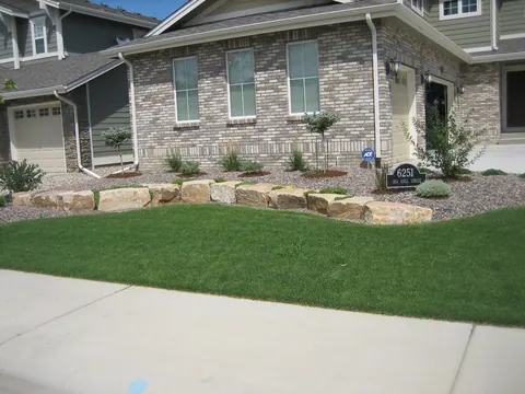 Low sandstone boulder wall along a gravel bed in front of a brick home and lawn.