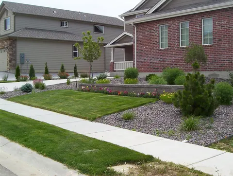 Low boulder wall and gravel bed with evergreens along a sidewalk in front of two homes.