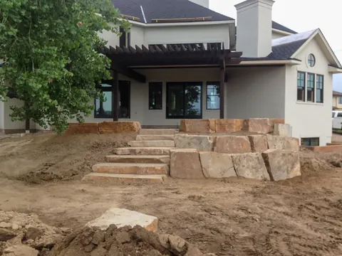 Stacked sandstone boulder wall with built-in steps rising toward a home with a pergola.