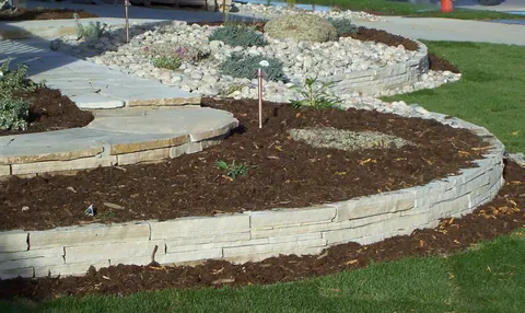 Curved stacked flagstone wall around a raised mulch bed with a flagstone path and river rock.