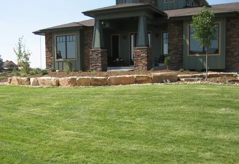 Row of sandstone boulders forming a low wall between a green lawn and a craftsman home.