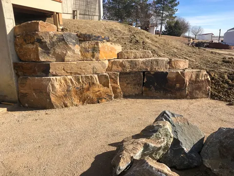 Stacked sandstone boulder retaining wall holding back a graded slope beside large dark boulders.