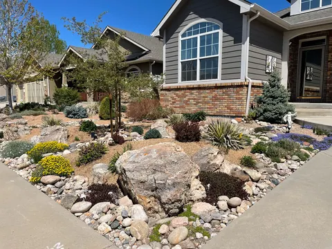 Corner lot xeriscape with a large boulder, spruce, purple flowers and river rock edging in Loveland.
