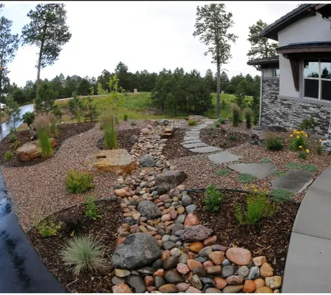 River rock dry stream bed beside flagstone stepping stones, grasses and pines near a stucco home.